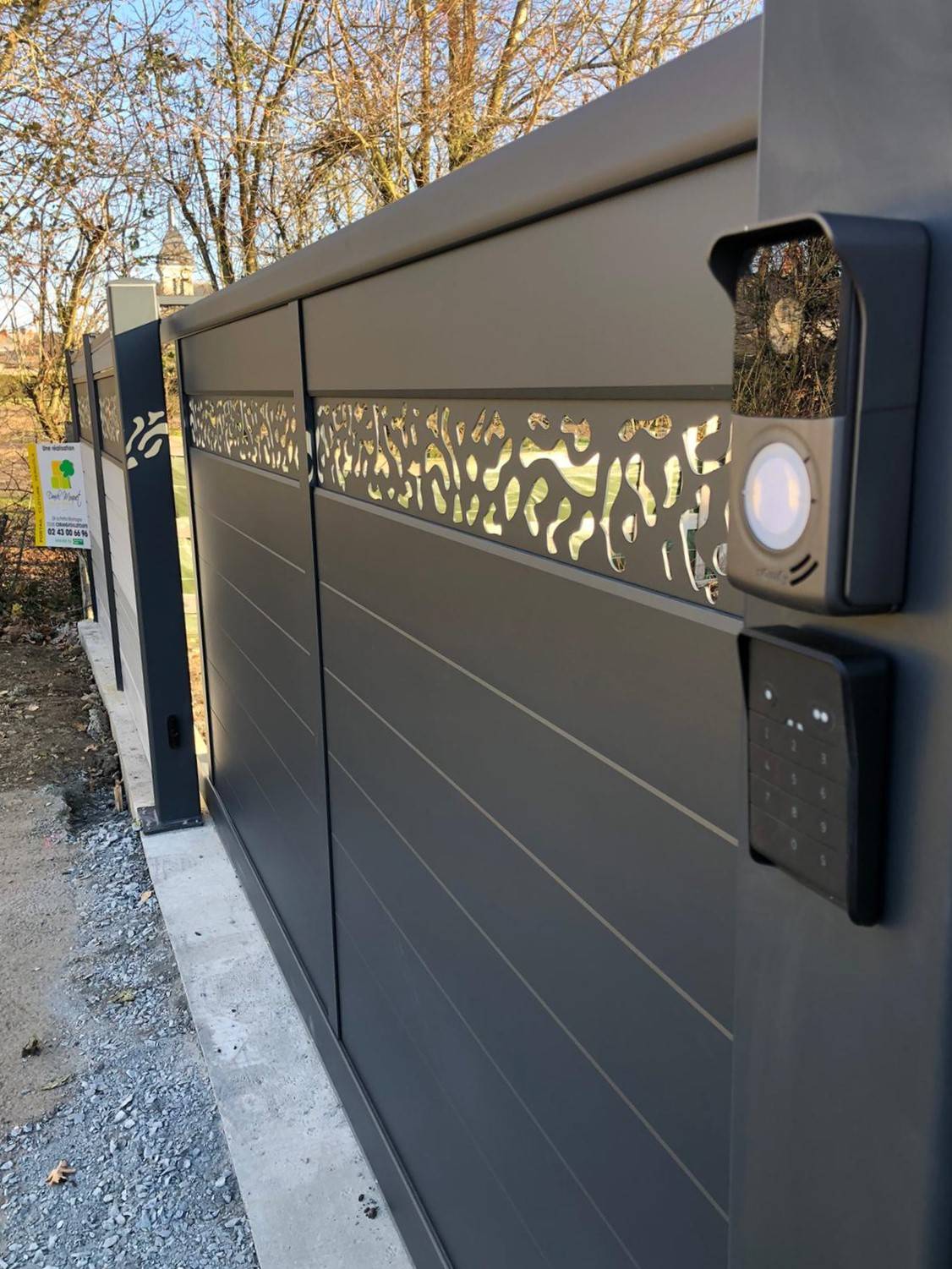 Portail en aluminium coulissant sur mesure et personnalisé avec un visiophone solaire et un clavier a code numérique, le portail posséde egalement un lame décorative en aluminium crée par découpe laser pour une réalisation original crée par Daniel Moquet 
